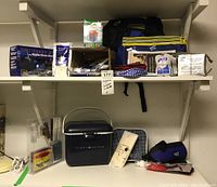 Full view of shelf showing all items in the lot including Tommy Hilfiger cooler, solar dynamo radio box, small bags, water bottle, hand sanitizer and packaged items.