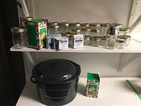 Photo of large black speckled enameled canning pot with lid and rack underneath a shelf holding multiple glass canning jars and two cardboard boxes of Bernardin snap lids, plus a clear glass shaker jar on the right.
