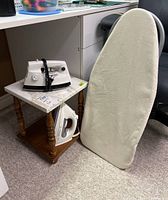 Proctor Silex and T-Fal irons, tabletop ironing board with white cover, wooden side table with stone top, all shown together on carpeted floor near office furniture.