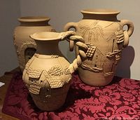 Three pottery pieces on a red patterned cloth: two vases and one jug, all featuring raised relief designs of houses and palm trees.