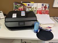 Front view of HP OfficeJet 4650 printer with assorted paper supplies behind including Avery labels and Office Depot photo paper.