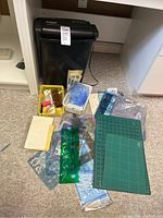 Photo showing Sylvania black paper shredder with various craft cutting items including cutting mats, plastic stencils, rulers, and small tools organized in two baskets on the floor.