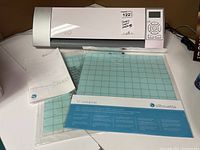 Front view of the Silhouette Cameo machine, two cutting mats still in packaging, manual beside mats, machine powered off.