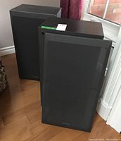 Two Technics floor speakers standing on hardwood floor near window, both covered with black fabric grills bearing Technics branding.