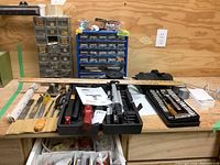 Full view of tools and hardware arranged on workbench including laser level tripod, ratchet set, chisels, rasps, files, knee pads, small containers of nuts and bolts, ruler.