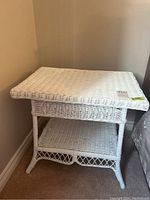 White wicker side table positioned in a corner with visible top and lower shelf, showing the wicker weave and curved front edge.