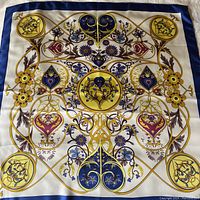 Top view of entire silk scarf showing intricate floral and heart-shaped pattern in gold, blue, and violet on white base with royal blue border.