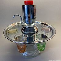 Front view of liquor dispenser with chrome rotating tray holding 4 shot glasses in yellow, amber, orange, green. Red pump button visible on top.