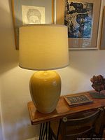 Full view of large ceramic lamp with mustard yellow rounded base and beige drum shade on wooden table, positioned against a wall with framed art.