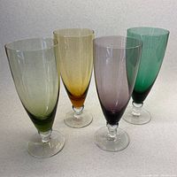 Four different colored mid-century modern blown glass stemmed champagne flutes standing upright side by side on a light surface.