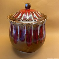 Front view of the mid-century modern lidded vessel showing the rounded body with red and chocolate drip glaze on cinnamon color base and the knobbed lid with scalloped edge.