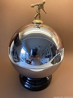 Front view showing highly polished chrome finish shaped like a bowling ball with a small bowler figurine handle on top reflecting room surroundings and person taking photo