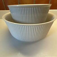 Photo showing two white round casserole dishes nested, highlighting their size difference and linear ribbed exterior pattern.