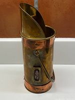 Tall cylindrical brass container with copper riveted bands, handle, and a match striking slot on the side