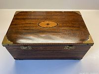 Top view of wooden box showing dark wood grain, oval inlay in center of lid, brass corner caps at all four corners.