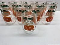 Front view of 10 identical vintage little juice glasses arranged closely, showing orange fruit and green leaves decoration on clear glass.