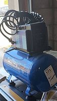 Blue horizontal tank air compressor with black motor case and coiled hose, on/off switch visible. Mastercraft brand and capacity label on tank.