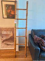 Full view of the bamboo ladder showing five rungs and vertical side rails, set against a wall beside framed Godzilla posters and a black leather couch.