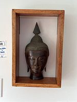 Front view of metal Buddha head in wooden shadow box shelf showing detailed facial features and wood grain.