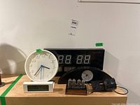 Five clocks including a white large analog clock, white alarm clock with LCD, black large segmented LED clock, black mantel clock with tubes and power adapter on shelf