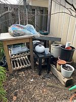 View showing outdoor setup of garden supplies including tables, stools, several ceramic and plastic planters, and a bag of yellow seed soil on the small wooden table.