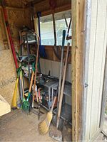 View of multiple garden tools including shovels, rakes, pitchfork, broom, watering cans, and hand tools arranged inside a shed corner.