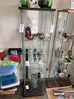 Full view of glass display cabinet showing glass sides, three shelves inside, wood base and top, with miscellaneous items on and beside it for scale.
