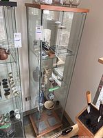 View of full glass cabinet showing three glass shelves and wood base and top with cabinet contents from adjacent lot visible.