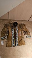 Full view of light brown buckskin coat with fringed sleeves and shoulders, showing hand beaded trim on front and cuffs.