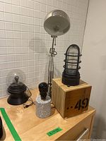 Four lamps on a wooden surface against a white tiled wall. Includes black skull lamp with terrazzo base, industrial cage lantern on wooden box marked '..49', metallic arc desk lamp, and black lamp with globe bulb cover.
