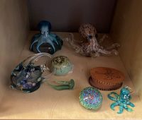 Various handmade marine figures including glass octopus, ceramic sea urchin, millefiori paperweight, glass fish, small octopus and wooden box on wooden shelf