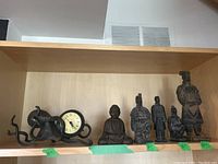 Seven black metal figures and one small clock displayed on a wood shelf, all items related to Asian-themed decor.