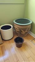 Three planters placed on a wooden floor against a light green wall; includes a large brown glazed pottery planter with floral and bird designs, medium dark brown pottery planter, and an off-white plastic planter.