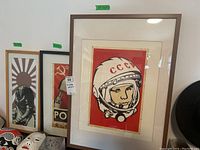 Three framed vintage posters leaning against a wall, featuring Soviet era imagery including a cosmonaut with CCCP helmet visible in front.