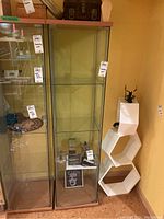 Tall glass shelving unit with three glass shelves and wooden top, shown next to matching shelving unit and white hexagonal shelves, with some decorative items inside and around it.