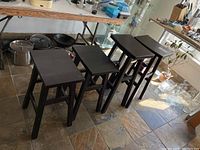 View of six wooden stools arranged in two rows under a wooden table, showing dark brown finish and rectangular seats.