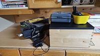 Photo showing Mastercraft steel vise, handcrafted wooden box, yellow container with Cobra walkie talkies, and gray mitre box on a wooden workbench.