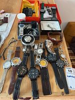 Overhead view of multiple analog watches laid out on wooden surface with spare straps in boxes and tin storage