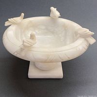 Front angle view of small white alabaster pedestal bowl with four carved birds on the rim and square base.