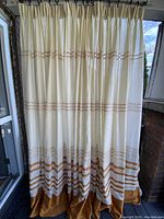 Four off-white drapery panels hanging, showing cinnamon colored border and woven cutout stripes, gathered tops, bottom hem solid cinnamon color.