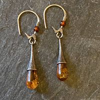 Pair of sterling silver drop earrings with cone-shaped settings holding amber drops and small amber beads on hooks, shown on dark textured background.