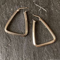 Pair of softly rounded triangular silver earrings laid on a black textured surface, showing the shape and clasp of the earrings.