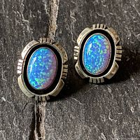 Pair of oval synthetic opal stud earrings set in serrated edge sterling silver bezels shown from above on a dark textured surface.