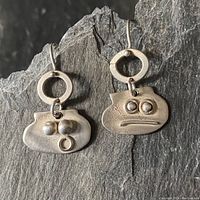 Pair of sterling silver earrings with small abstract face designs, showing overall view and drop length on a dark stone background.