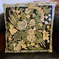 Front view of the embroidered pillow on a black chair showing the floral green and cream design on a black background.