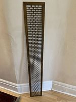 Full view of the long narrow rectangular brass grate standing upright against a corner wall showing overall size and shape.