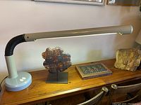 Full view of the lamp on a wooden desk showing the weighted pale grey metal base, the black flexible gooseneck, and the long taupe synthetic shade.