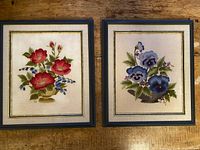 Both framed embroidered floral artworks shown side by side on a wooden surface. One depicts red flowers and the other blue pansies. Each is bordered by a double-line gold decoration under glass.