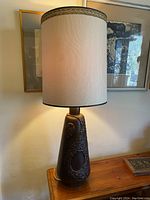 Full view of the lamp on a wooden surface with art framed on the wall in the background. Lamp is lit and shows its height and overall form.