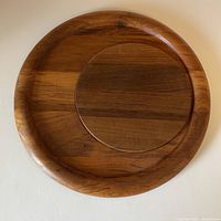 Full top-down view of the circular teak cheeseboard with separate round insert fitted inside. Shows the slightly raised edge of the cheeseboard and the grain pattern of the wood.